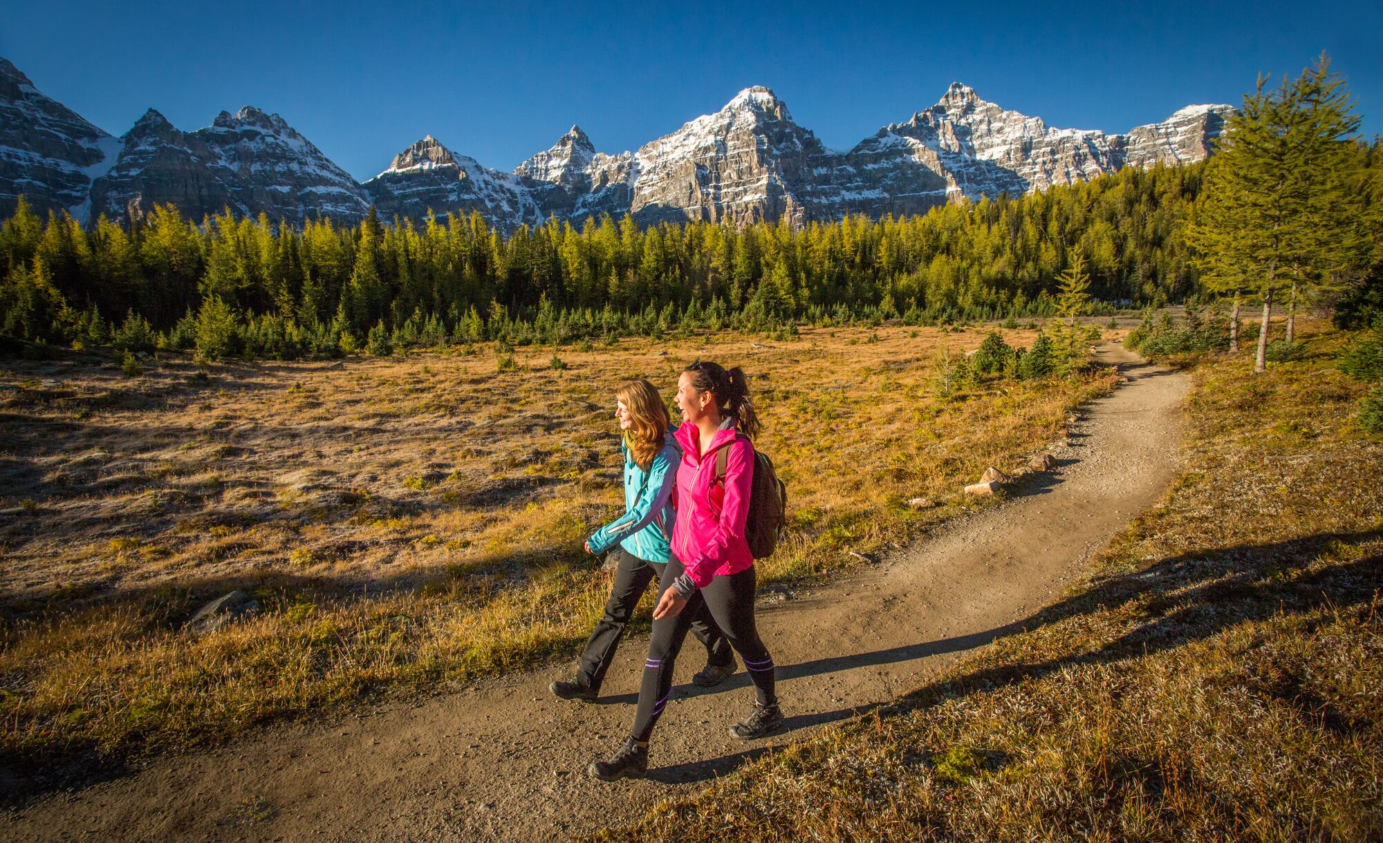 Two women walk in Larch Valley with mountain peaks behind them in Banff National Park. 